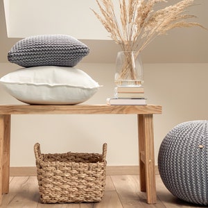 May include: A wooden bench with a woven basket, a gray knitted pouf, and two gray knitted pillows. A vase with dried pampas grass sits on top of a stack of books on the bench.