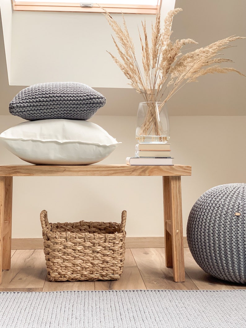 May include: A wooden bench with a woven basket, a gray knitted pouf, and two gray knitted pillows. A vase with dried pampas grass sits on top of a stack of books on the bench.