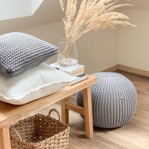 May include: A wooden bench with two gray knitted pillows, a white pillow, and a woven basket. A vase with dried pampas grass sits on top of a stack of books. A gray knitted pouf sits on the floor next to the bench.