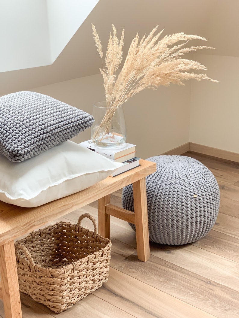 May include: A wooden bench with two gray knitted pillows, a white pillow, and a woven basket. A vase with dried pampas grass sits on top of a stack of books. A gray knitted pouf sits on the floor next to the bench.