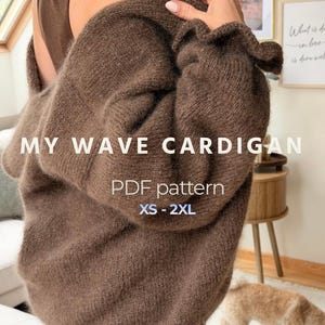 May include: A brown knit cardigan with long sleeves and a ruffled cuff detail. The text MY WAVE CARDIGAN PDF pattern XS - 2XL is overlaid on the image. The garment is worn over a brown tank top, showcasing a soft, fuzzy texture and a loose fit.
