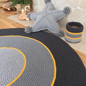 May include: A round, grey and yellow knitted rug with a smaller grey and yellow knitted basket. A grey knitted star-shaped cushion with pom-poms is in the background.