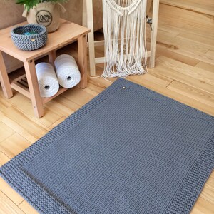 May include: A gray knitted rug with a thick border. The rug is on a wooden floor.