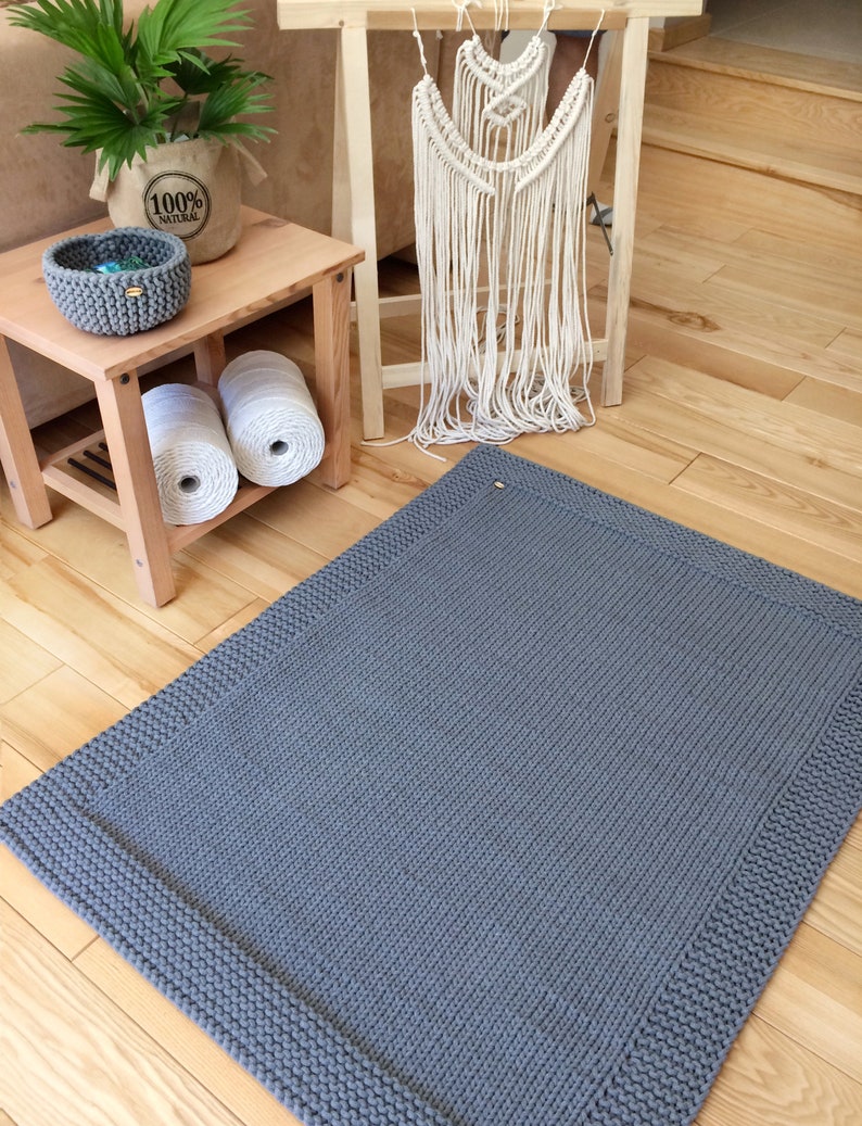May include: A gray knitted rug with a thick border. The rug is on a wooden floor.