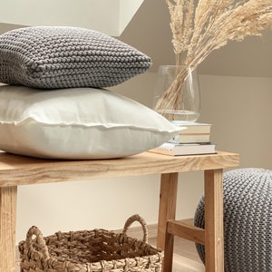 May include: A wooden bench with a gray knitted ottoman, a woven basket, and two pillows. One pillow is gray and the other is white. A vase with dried pampas grass sits on the bench.