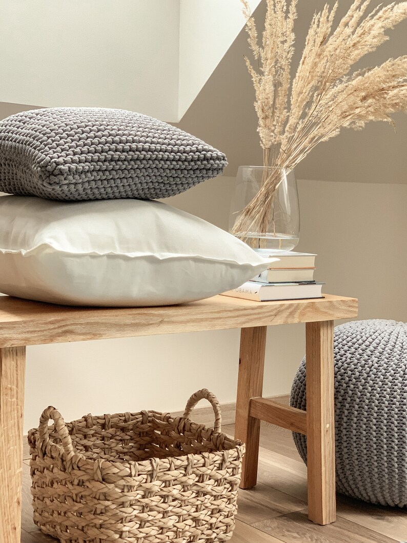 May include: A wooden bench with a gray knitted ottoman, a woven basket, and two pillows. One pillow is gray and the other is white. A vase with dried pampas grass sits on the bench.