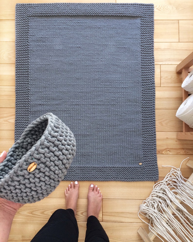 May include: A gray knitted rug with a thick border. The rug is on a wooden floor. A gray knitted basket is in the foreground.