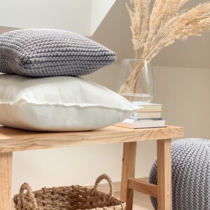 May include: A wooden bench with a gray knitted pouf, a woven basket, and two gray knitted pillows. The bench is in front of a white wall with a vase of dried pampas grass.