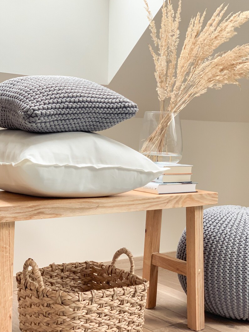 May include: A wooden bench with a gray knitted pouf, a woven basket, and two gray knitted pillows. The bench is in front of a white wall with a vase of dried pampas grass.