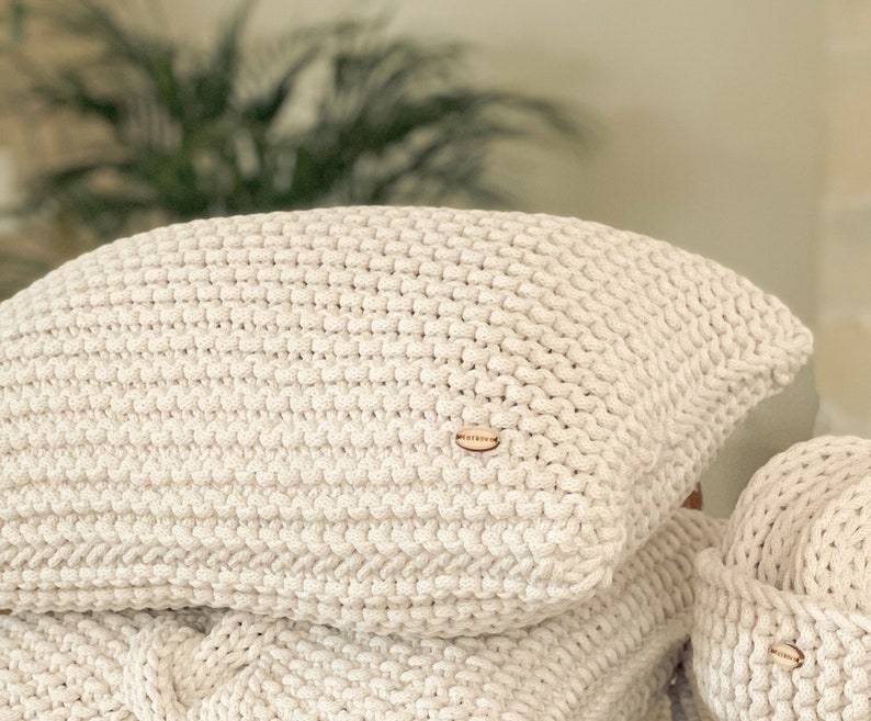 May include: A close-up of a cream-colored knitted throw pillow with a small, natural shell button in the center.
