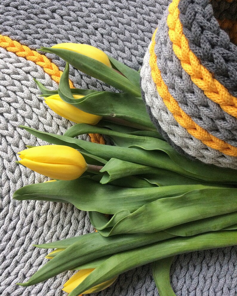 May include: A bunch of yellow tulips with green leaves on a grey knitted rug with yellow stripes.