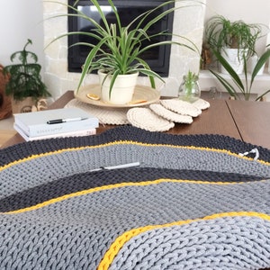 May include: A grey and black crocheted rug with yellow accents. The rug is on a wooden table with a plant and a book on it.