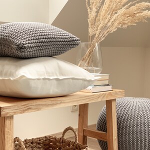 May include: A wooden bench with a gray knitted ottoman, a woven basket, and two gray knitted pillows. A vase with dried pampas grass sits on top of a stack of books.