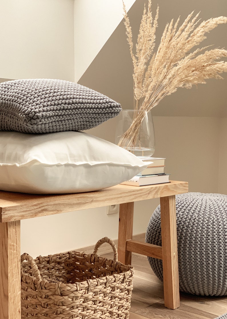 May include: A wooden bench with a gray knitted ottoman, a woven basket, and two gray knitted pillows. A vase with dried pampas grass sits on top of a stack of books.
