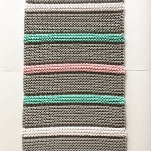 Puede incluir: Una manta rectangular tejida a mano con rayas horizontales. La manta presenta bandas alternas de gris, blanco, rosa claro y verde menta. Los bordes superior e inferior tienen un dise&ntilde;o acanalado. Se adjunta una peque&ntilde;a etiqueta redonda de madera.