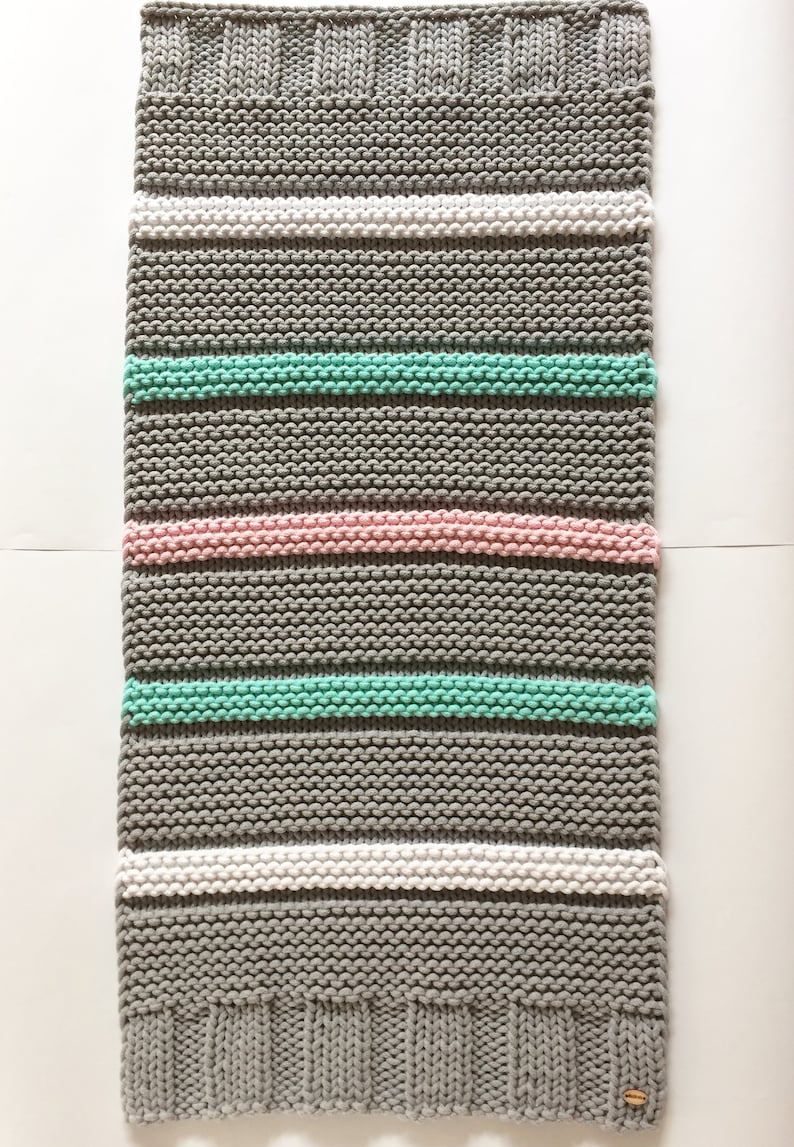 Puede incluir: Una manta rectangular tejida a mano con rayas horizontales. La manta presenta bandas alternas de gris, blanco, rosa claro y verde menta. Los bordes superior e inferior tienen un dise&ntilde;o acanalado. Se adjunta una peque&ntilde;a etiqueta redonda de madera.