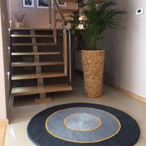May include: A round, grey and black crocheted rug with a yellow border. The rug is in a hallway with a wooden staircase and a potted plant.
