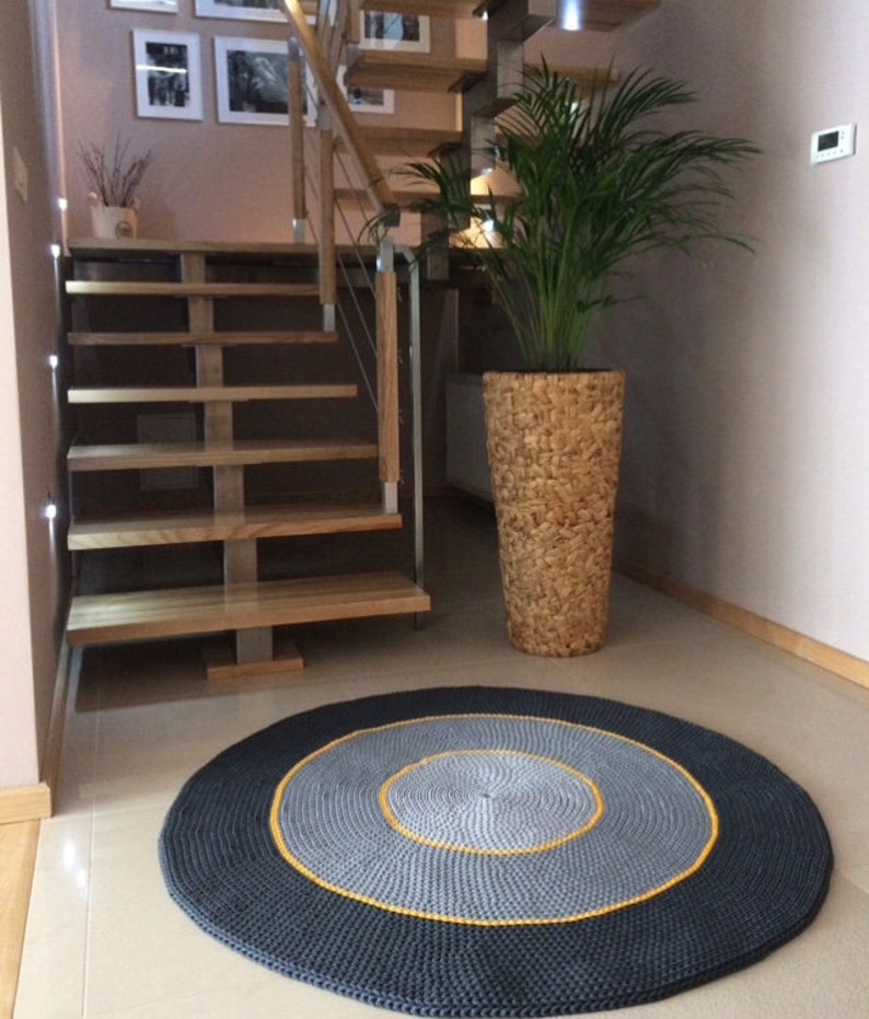 May include: A round, grey and black crocheted rug with a yellow border. The rug is in a hallway with a wooden staircase and a potted plant.