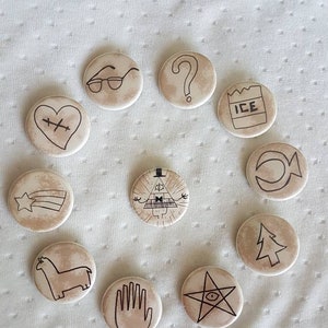 May include: A set of 11 round pins with various designs from the TV show Gravity Falls. The pins feature characters and symbols from the show, including a triangle with an eye, a shooting star, a llama, a hand, a tree, a question mark, a heart with a bandage, a crescent moon, a pair of glasses, a box of ice, and a star with an eye.