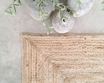 Puede incluir: Una alfombra de yute natural con un diseño tejido y un borde grueso. La alfombra es de color marrón claro y tiene una sensación natural y terrosa.