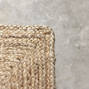 May include: Close-up of a natural fiber rug with a woven texture. The rug is a light beige color and has a square shape. The rug is on a gray concrete floor.