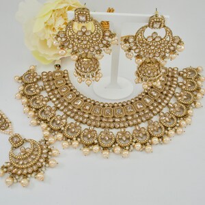 May include: A gold-toned jewelry set featuring a necklace, earrings, and a headpiece. The set is adorned with clear and pearl-like accents. The earrings are large, and the necklace has a detailed, ornate design. The headpiece is delicate and complements the set.