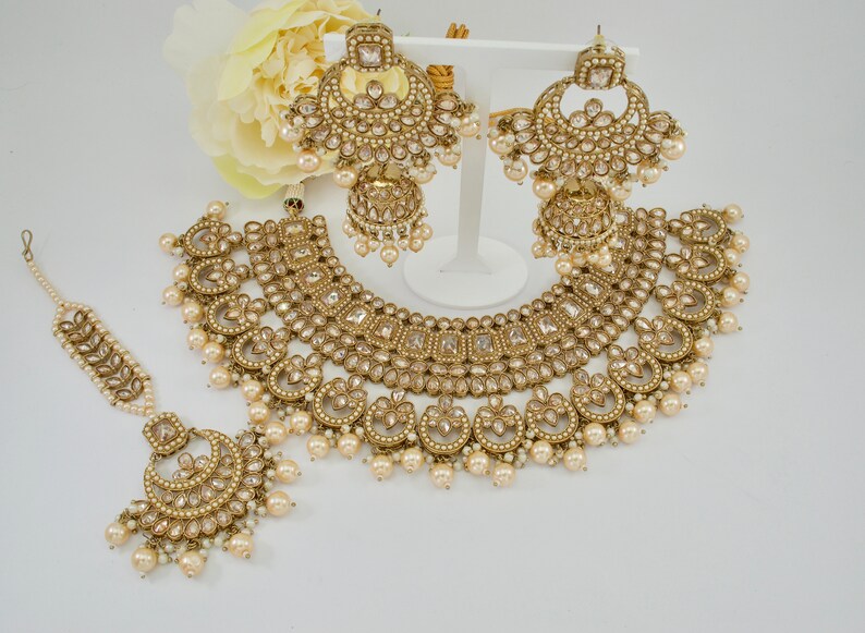 May include: A gold-toned jewelry set featuring a necklace, earrings, and a headpiece. The set is adorned with clear and pearl-like accents. The earrings are large, and the necklace has a detailed, ornate design. The headpiece is delicate and complements the set.