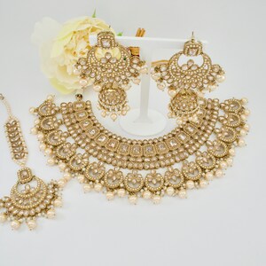 May include: A gold-toned bridal jewelry set featuring a necklace, earrings, and a headpiece. The set is adorned with clear crystals and faux pearls. The necklace and earrings have a layered design, and the headpiece is delicate. The jewelry is displayed on a white surface.
