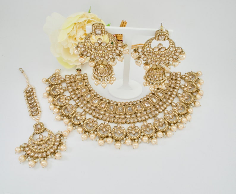 May include: A gold-toned bridal jewelry set featuring a necklace, earrings, and a headpiece. The set is adorned with clear crystals and faux pearls. The necklace and earrings have a layered design, and the headpiece is delicate. The jewelry is displayed on a white surface.