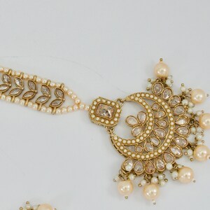 May include: An ornate gold-toned headpiece with a crescent moon design, adorned with clear crystals and faux pearls. A delicate strand of pearls connects to a section with leaf-shaped accents. The jewelry is likely for a special occasion.