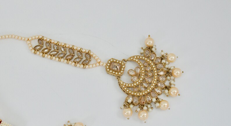 May include: An ornate gold-toned headpiece with a crescent moon design, adorned with clear crystals and faux pearls. A delicate strand of pearls connects to a section with leaf-shaped accents. The jewelry is likely for a special occasion.