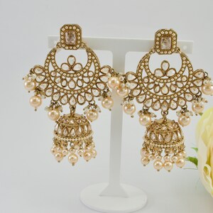 May include: Gold-toned chandelier earrings with a square top and a half-moon design. The earrings feature clear, teardrop-shaped accents and dangling faux pearls. A bell-shaped charm with more pearls hangs below.