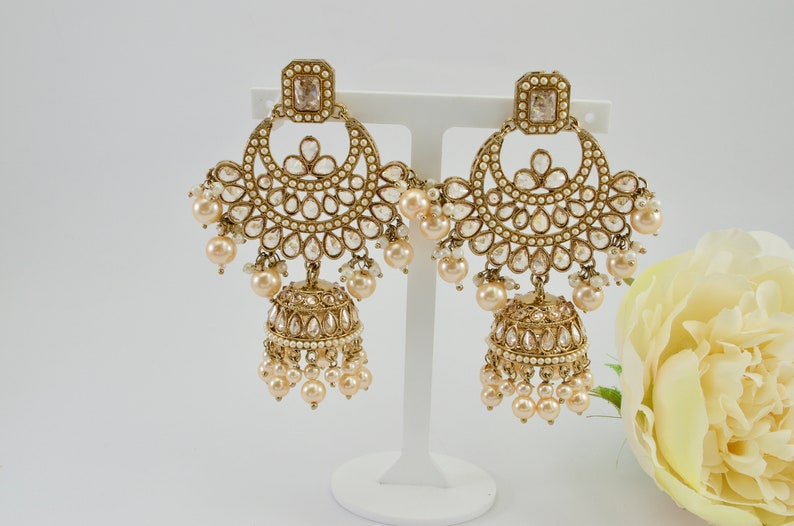 May include: Gold-toned chandelier earrings with a square top and a half-moon design. The earrings feature clear, teardrop-shaped accents and dangling faux pearls. A bell-shaped charm with more pearls hangs below.