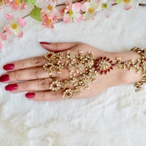 Peut inclure: Une chaîne de main dorée ornée de pierres précieuses rouges et transparentes et de petites perles blanches. Le bijou est porté sur une main avec du vernis à ongles rouge. Des fleurs roses sont en arrière-plan.