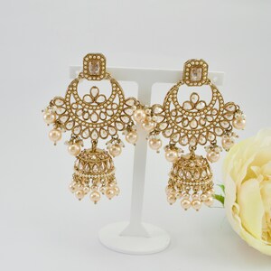 May include: A pair of gold-toned chandelier earrings with a square top, a half-moon design, and a bell-shaped bottom. The earrings are adorned with clear crystals and small, round, cream-colored pearls. The earrings are displayed on a white stand.
