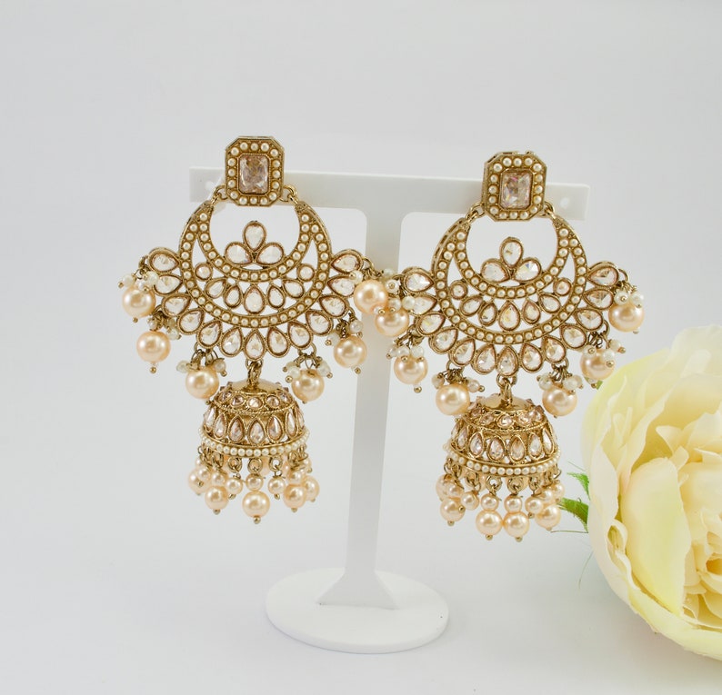 May include: A pair of gold-toned chandelier earrings with a square top, a half-moon design, and a bell-shaped bottom. The earrings are adorned with clear crystals and small, round, cream-colored pearls. The earrings are displayed on a white stand.