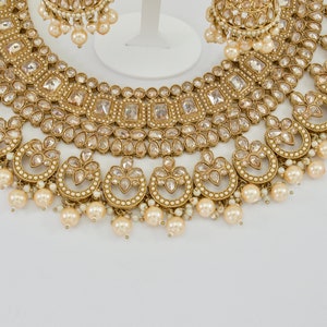 May include: A gold-toned bridal jewelry set featuring a necklace, earrings, and a bracelet. The necklace and earrings have clear, square-cut stones and dangling faux pearls. The jewelry is set against a white background.