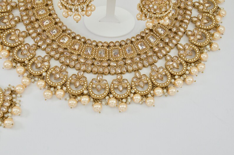 May include: A gold-toned bridal jewelry set featuring a necklace, earrings, and a bracelet. The necklace and earrings have clear, square-cut stones and dangling faux pearls. The jewelry is set against a white background.