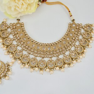 May include: A gold-toned bridal jewelry set featuring a necklace, a headpiece, and a tikka. The necklace has a curved design with clear stones and dangling pearls. The headpiece and tikka are adorned with similar details. A cream-colored flower is in the background.