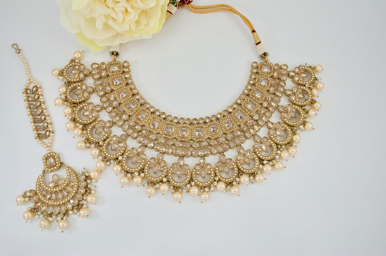May include: A gold-toned bridal jewelry set featuring a necklace, a headpiece, and a tikka. The necklace has a curved design with clear stones and dangling pearls. The headpiece and tikka are adorned with similar details. A cream-colored flower is in the background.