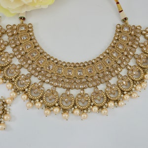 May include: An ornate gold-toned jewelry set featuring a necklace, a headpiece, and a pendant. The necklace has multiple layers of square and circular designs, adorned with clear stones and faux pearls. The headpiece and pendant mirror the necklace's design.