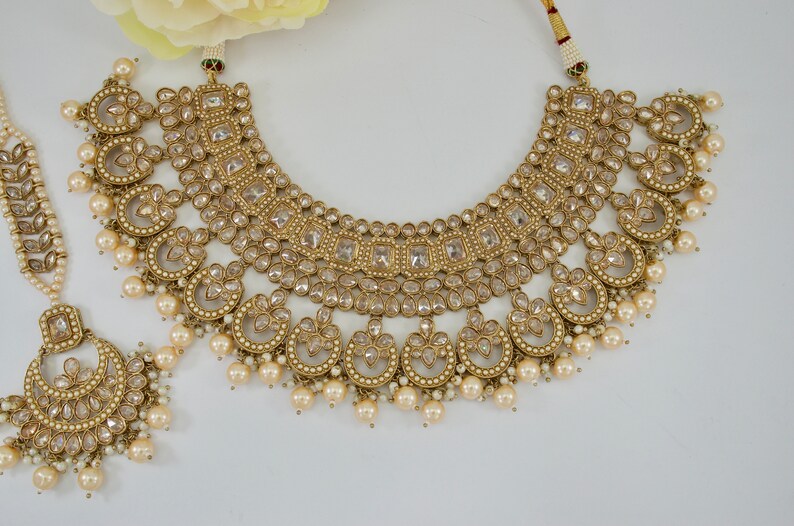 May include: An ornate gold-toned jewelry set featuring a necklace, a headpiece, and a pendant. The necklace has multiple layers of square and circular designs, adorned with clear stones and faux pearls. The headpiece and pendant mirror the necklace's design.