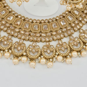 May include: A gold-toned jewelry set featuring a necklace and earrings. The necklace has a layered design with clear, square and teardrop-shaped stones, and dangling faux pearls. The earrings mirror the necklace's design, with pearl drops.