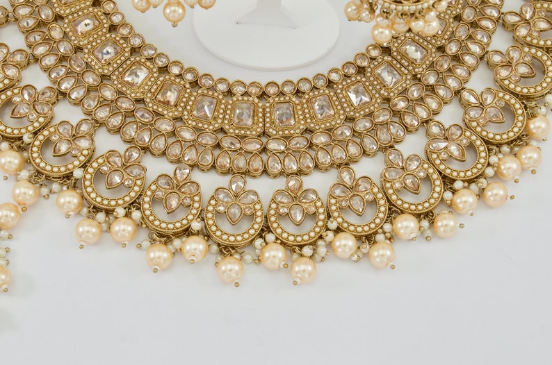 May include: A gold-toned jewelry set featuring a necklace and earrings. The necklace has a layered design with clear, square and teardrop-shaped stones, and dangling faux pearls. The earrings mirror the necklace's design, with pearl drops.