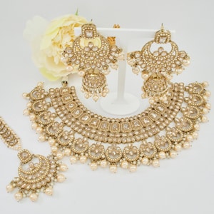 May include: A gold-toned jewelry set featuring a necklace, earrings, and a headpiece. The set includes clear and pearl-like accents. The earrings are displayed on a white stand. The necklace and headpiece have a matching design.