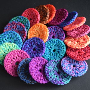 May include: A collection of colorful crocheted scrubbies in various shades of blue, green, red, orange, purple, and pink. The scrubbies are arranged in a circular pattern, with the largest scrubby in the center and the smallest scrubbies on the outside.