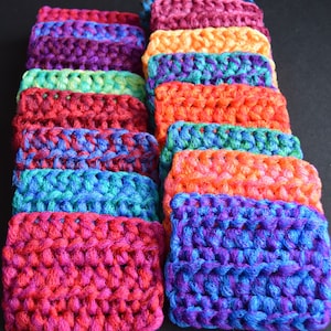 May include: A stack of colorful crocheted dishcloths in various shades of red, orange, yellow, green, blue, and purple. The cloths are made of a textured yarn and are perfect for cleaning dishes or scrubbing surfaces.