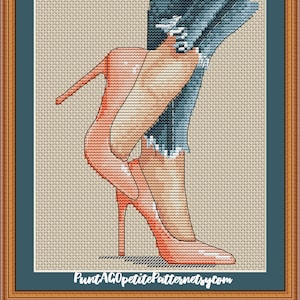 May include: Cross-stitch artwork featuring a pair of coral-colored high-heeled shoes and distressed blue jeans. The artwork is framed in a brown wooden frame with a teal mat. The text "PuntAGOpettitePatternetsy.com" is at the bottom.