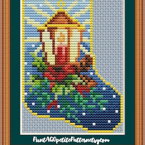 Könnte beinhalten: Ein Kreuzstichmuster einer Weihnachtsstrumpf mit einer Laterne und Stechpalme. Der Strumpf ist blau mit weißen Schneeflocken und die Laterne ist braun mit einer roten Kerze darin. Das Muster ist auf weißem Hintergrund mit dem Text "PuntAGOpetitePatternsetsy.com".