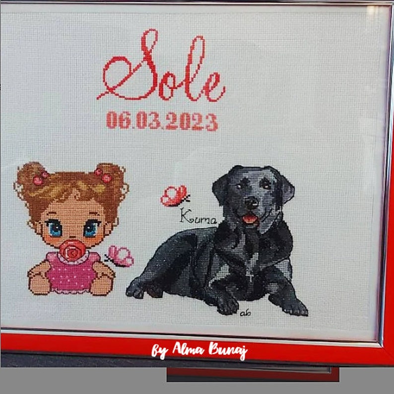 May include: Cross-stitch embroidery with a baby girl, a black Labrador Retriever, and the text "Sole 06.03.2023" in red. The embroidery is framed in a red and silver frame. The text "by Alma Bunaj" is at the bottom of the frame.
